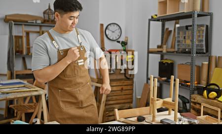 Un giovane asiatico in un'officina che indossa un grembiule marrone che esamina gli strumenti per la lavorazione del legno tra vari progetti di falegnameria. Foto Stock