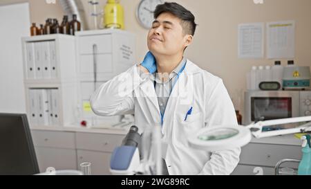 Un giovane asiatico in un camice bianco da laboratorio appare stanco o stressato in un ambiente di laboratorio clinico. Foto Stock