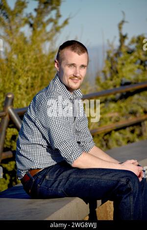 Il visitatore siede su una panchina di cemento in cima al Pilot Butte in Oregon. Indossa una camicia con abito a quadri e jeans blu. Sta sorridendo. Foto Stock