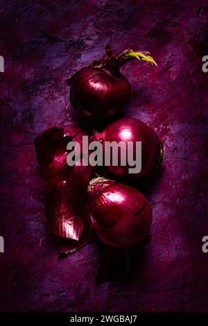 Cipolle rosse fresche su uno sfondo ruvido con sfumature di rosso e viola, vista dall'alto. Natura morta Foto Stock