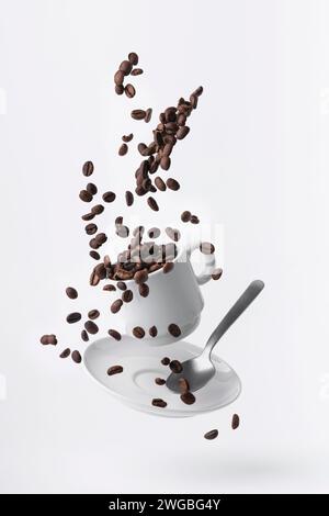 chicchi di caffè tostati versati da una tazza galleggiante e da un cucchiaino isolato su sfondo bianco Foto Stock