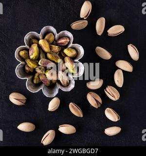 vista dall'alto su sfondo nero testurizzato, ciotola con semi di pistacchio sgusciati; sullo sfondo pistacchi in guscio. Foto Stock