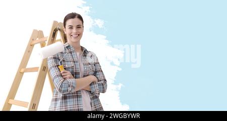 Giovane donna sorridente che dipinge la sua casa, si trova accanto a una scala e tiene in mano un rullo di pittura Foto Stock