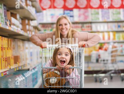 Ragazza allegra e carina seduta in un carrello al supermercato, sua madre la sta spingendo: Shopping di generi alimentari e concetto di stile di vita Foto Stock