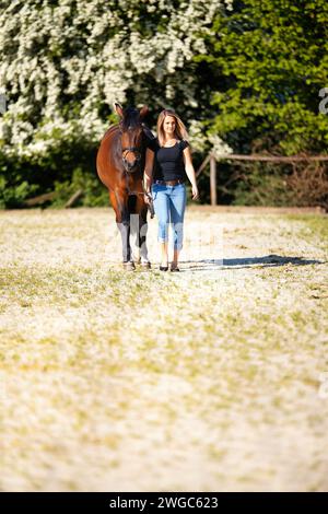 Una giovane donna sta con il suo cavallo su un'arena illuminata dal sole. Sullo sfondo, alberi in fiore e natura verde, modellano isolato di fronte alla sfocatura, fratello Foto Stock