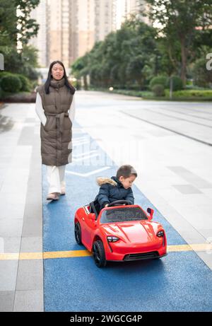 Un giovane bimbo multirazziale naviga con gioia sulla pista da corsa del quartiere a bordo di un'auto giocattolo sportiva rossa, sorridendo e assumendo il controllo di Foto Stock