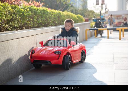Un giovane bimbo multirazziale naviga con gioia lungo la strada del quartiere a bordo di un'auto giocattolo sportiva rossa, sorridendo e assumendo il controllo della CA giocattolo Foto Stock