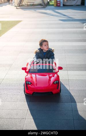 Un giovane bimbo multirazziale naviga con gioia lungo la strada del quartiere a bordo di un'auto giocattolo sportiva rossa, sorridendo e assumendo il controllo della CA giocattolo Foto Stock