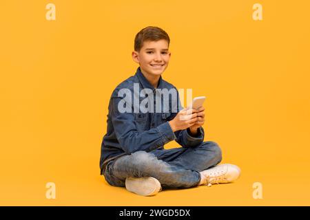 Adolescente sorridente con smartphone in mano seduto a gambe incrociate su sfondo giallo Foto Stock