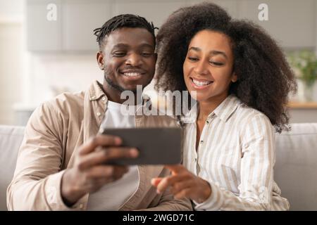 Allegra coppia Millennial nera che utilizza lo smartphone per realizzare Selfie a casa Foto Stock