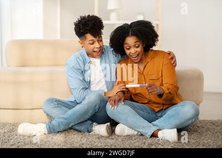 Una giovane coppia felice si siede sul pavimento con un test di gravidanza positivo Foto Stock