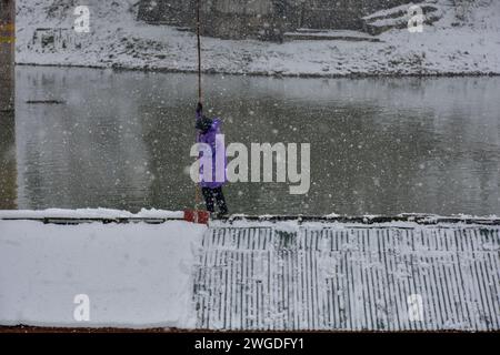 Un uomo libera la neve dal tetto della sua casa galleggiante durante una forte nevicata a Srinagar. La valle del Kashmir è stata testimone di nevicate fresche che coprono diverse parti, tra cui la capitale Srinagar la domenica, interrompendo il movimento stradale. La nevicata fresca è stata una sorpresa dopo un Chillai Kalan insolitamente secco e quasi senza neve, il tradizionale periodo di 40 giorni di rigido inverno. Foto Stock