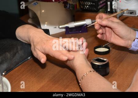 Il manicurista tiene le dita del cliente in mano e applica una vernice incolore alle unghie con un pennello sottile. Foto Stock