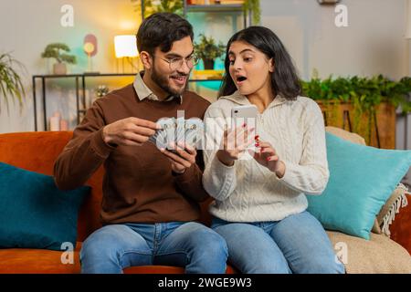 Felice coppia indiana con soldi in contanti e pianificazione smartphone discutendo insieme di investimenti seduti sul divano nel soggiorno di casa. L'uscita di successo ha stupito una famiglia diversificata sul divano dell'appartamento. Foto Stock
