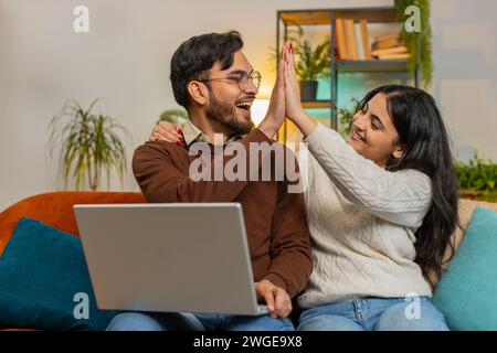 Allegra giovane e variegata coppia in abiti casual con laptop e sorpresa con la lotteria vincente per e-mail sul divano nel soggiorno di casa. La famiglia indiana felice dà cinque pugni stringenti in appartamento. Foto Stock