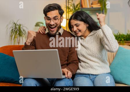Allegra giovane e variegata coppia in abiti casual che usa il computer portatile sorpreso con il jackpot di gioco online della lotteria che ha vinto per e-mail sul divano nel soggiorno di casa. Felice famiglia ispanica che stringe pugni in appartamento Foto Stock