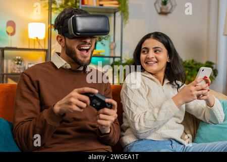 Uomo felice che esce con occhiali VR che giocano a videogame con joystick seduto accanto alla ragazza indiana che usa lo smartphone seduto sul divano nel soggiorno di casa. Famiglia felice che trascorre il fine settimana in appartamento Foto Stock