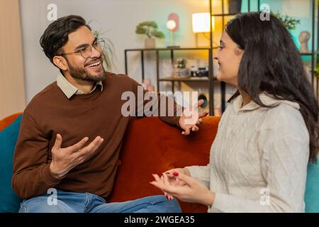 Sorridente giovane coppia di famiglia in abiti informali seduti sul divano insieme e facendo una bella conversazione nel soggiorno di casa. Uomo ispanico che parla con la ragazza indiana che si rilassa sul divano dell'appartamento. Foto Stock