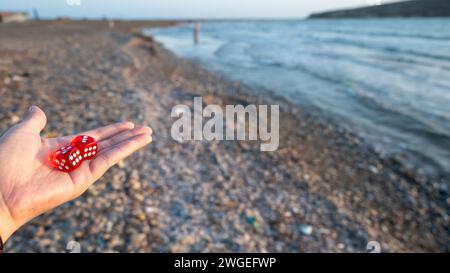 La mano di una donna con i dadi rossi sullo sfondo di una spiaggia. Foto Stock