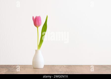 Piccolo tulipano rosa singolo con foglia verde in vaso bianco su un tavolo di legno con sfondo bianco Foto Stock