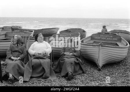 Brighton Beach anni '1970 Regno Unito. Tre vecchi vacanzieri, avvolti da coperte, siedono su sdraio mantenendosi al caldo sulla spiaggia di Brighton, riparandosi in parte dal vento dietro le barche da pesca del Deep Sea Anglers Club. Brighton, East Sussex, Inghilterra 1971. HOMER SYKES Foto Stock