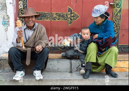 Un uomo con una ruota tibetana si siede su gradini di pietra con sua moglie e suo figlio in Piazza Barkhor, Lhasa. Foto Stock