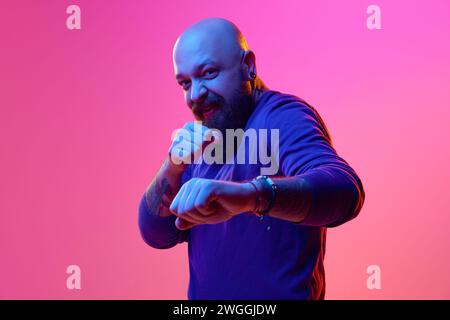 Divertente lotta. Ritratto in gel di un uomo calvo barbuto artistico con maglione blu che posa emotivamente su sfondo rosa alla luce del neon Foto Stock