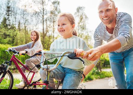 Sorridente padre con due figlie durante una passeggiata all'aperto. Insegna alla ragazza più giovane a fare una bicicletta. Amano stare insieme nel parco estivo della città. Ha Foto Stock