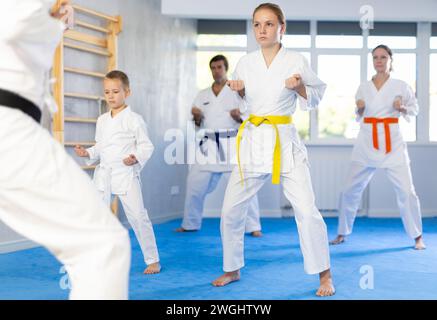 Teen girl con la sua famiglia in kimono e cinture colorate che praticano karate con pugni durante le lezioni di arti marziali di gruppo in palestra, accompagnata da un allenatore Foto Stock