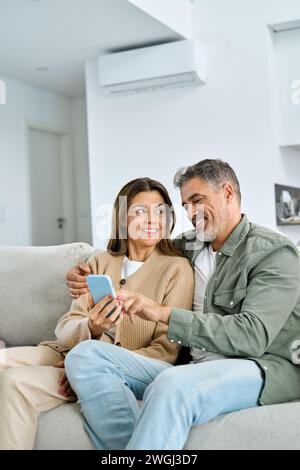 Una coppia di mezza età felice che usa il telefono seduto a casa e si rilassa sul divano. Verticale. Foto Stock