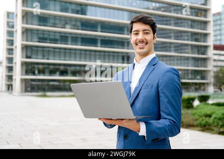 Un uomo d'affari ispanico sorride mentre lavora al suo laptop, simboleggiando l'intersezione tra il business tradizionale e la mobilità moderna con un grattacielo sullo sfondo Foto Stock