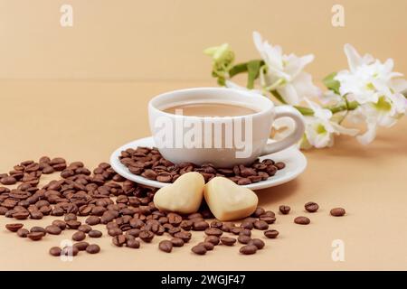 Tazza di caffè e chicchi di caffè, caramelle a forma di cuore e fiori bianchi su sfondo beige. Il giorno di San Valentino. Primo piano. Foto Stock