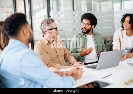 I colleghi conversano intorno a un notebook, con un membro che spiega un concetto, l'attenzione del team e il linguaggio del corpo aperto indicano un ambiente di lavoro collaborativo e ricettivo Foto Stock