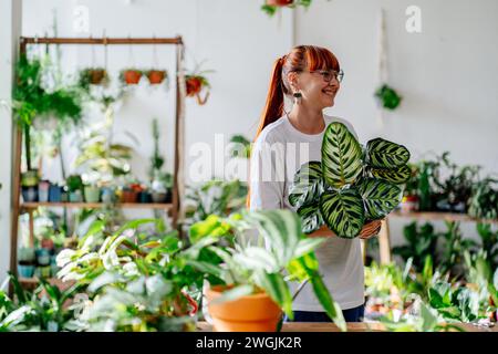 Donna giardiniere in tuta arancione che innaffia piante in vaso in serra circondata da piante e vasi, utilizzando lattine bianche in metallo. Casa Foto Stock