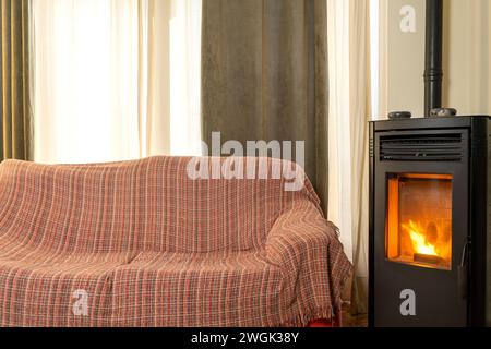 Soggiorno con comodo divano da indossare con copertura. Stufa a pellet in ghisa nera a casa. Impianto di riscaldamento ecologico. Pellet moderno Foto Stock