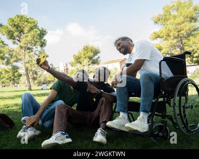 Gruppo di giovani amici maschi diversi uno di loro su una sedia a rotelle che si fa un selfie in un parco Foto Stock