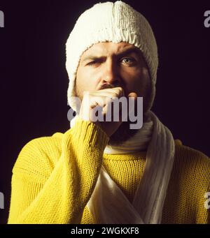 Uomo malvagio con influenza o polmonite che tossisce nel pugno. L'uomo malato ha la tosse come sintomo di influenza, raffreddore o bronchite. Uomo malato in maglione e cappello Foto Stock