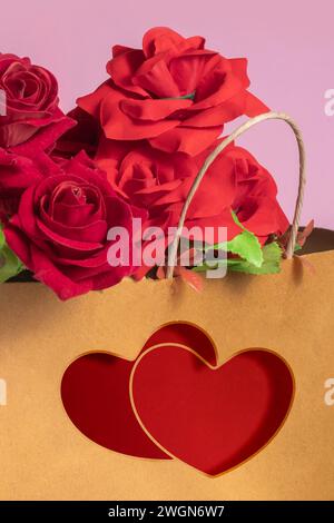 Rose rosse all'interno di una borsa della spesa in carta marrone con due cuori rossi, borsa regalo stampata su sfondo rosa Foto Stock