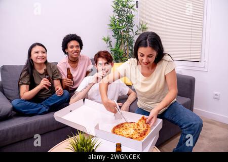 Un gruppo di quattro amici si gusta pizza e birre in un comodo soggiorno con una pianta di casa. Foto Stock