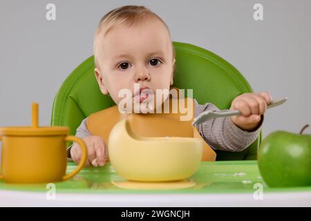 Piccolo bambino carino che mangia cibo sano in seggiolone su sfondo grigio Foto Stock