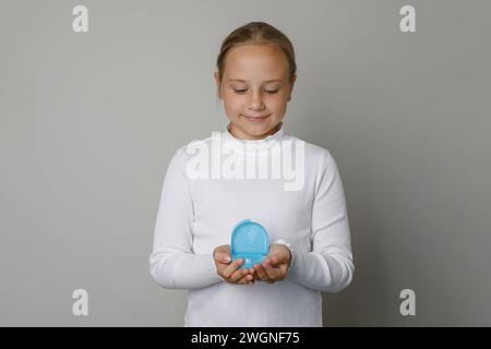 La bambina tiene tra le mani la scarpa in silicone per l'allineamento dei denti sullo sfondo grigio della parete Foto Stock