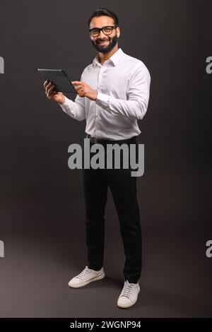 Un allegro uomo d'affari indiano con una camicia bianca interagisce con un tablet digitale, la sua posizione a tutto il corpo e il sorriso coinvolgente che trasmette accessibilità e competenza tecnologica Foto Stock