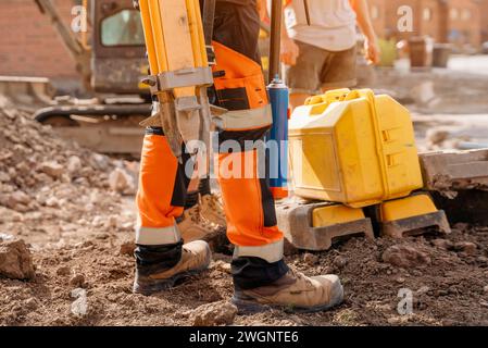 Primo piano sulle scarpe di sicurezza di un geometra ingegnere civile in piedi sul cantiere con il suo treppiede e lo strumento Foto Stock