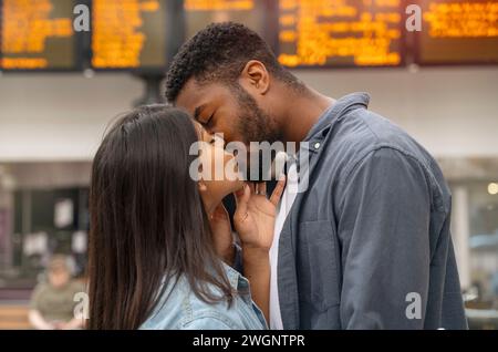 una giovane coppia di famiglie alla stazione ferroviaria si baciava davanti agli orari Foto Stock