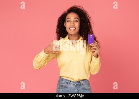 Felice donna nera con gesti con carta di credito su sfondo rosa Foto Stock