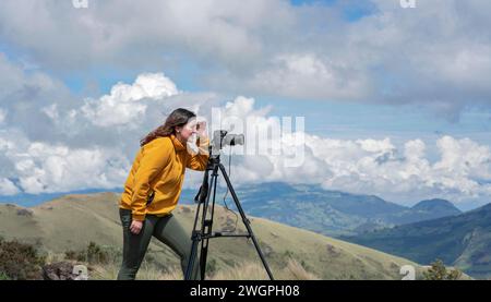 Bella giovane donna latinoamericana con fotocamera e cavalletto che scatta foto di paesaggi sulla cima di una montagna durante una giornata di sole Foto Stock