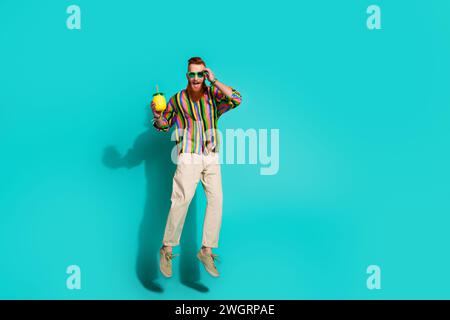 Foto a grandezza naturale di un uomo di successo che salta con occhiali da sole tropicali cocktail touch sembra fresco isolato su sfondo di colore ciano Foto Stock