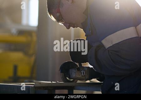 Il meccanico dei metalli lavora con la smerigliatrice angolare in officina. Luce cinematografica retroilluminata. Foto. Ritratto di un vero uomo al lavoro. Macina il metallo. Foto Stock