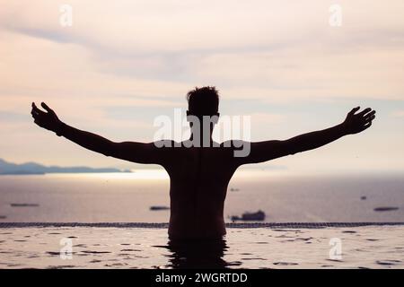 Silhouette di un giovane ragazzo felice con le braccia allungate nella piscina a sfioro, godendosi la vista al tramonto. Foto Stock