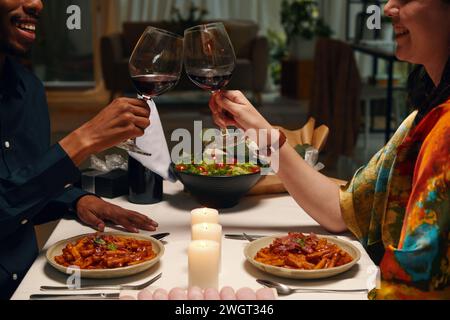 Bicchierino ritagliato di giovani valentine interculturali felici che brulicano con bicchieri di vino rosso mentre si siede al tavolo servito con cibo fatto in casa Foto Stock